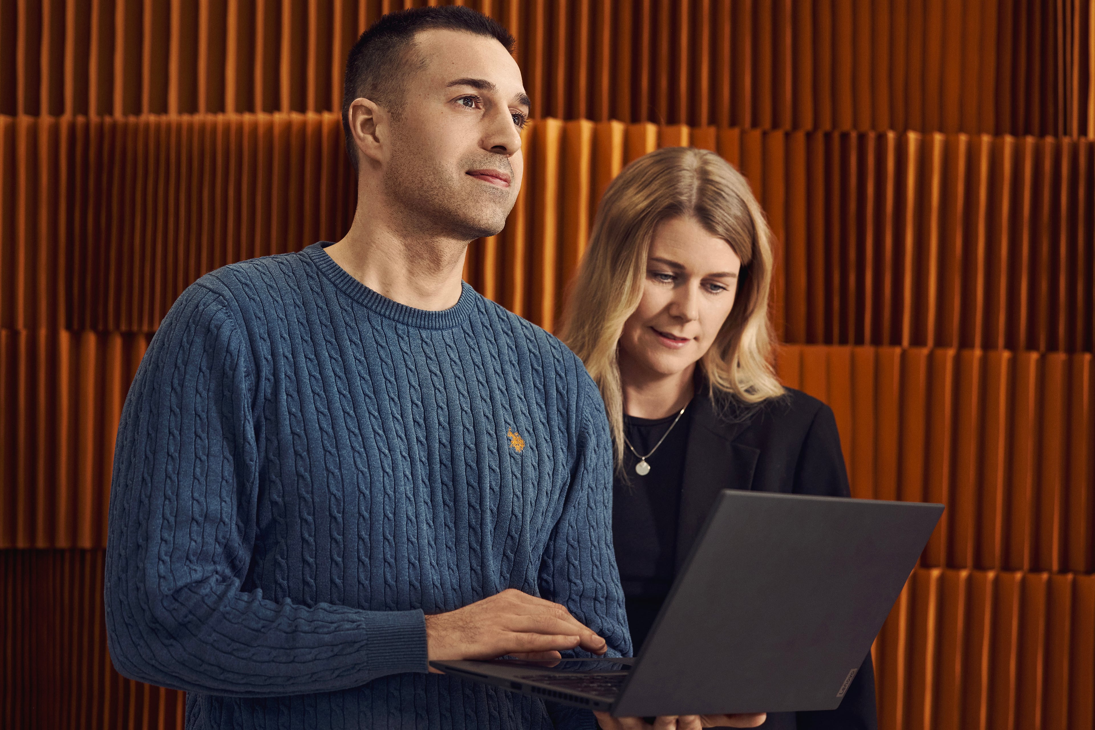 Two people working together at a laptop in front of an orange ribbed wall panel