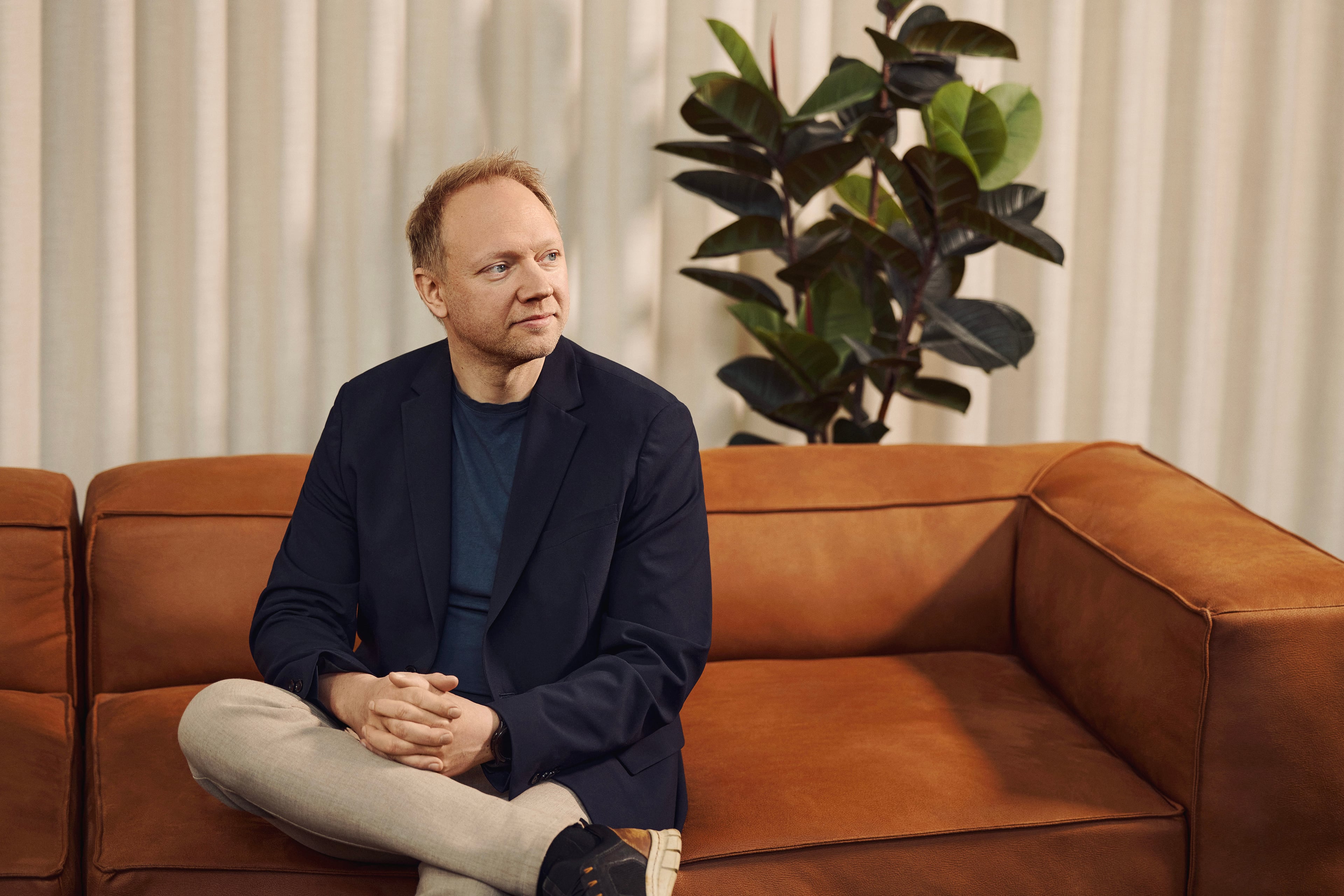 Man in navy blazer sitting on cognac-colored sofa in front of white curtains and green plant