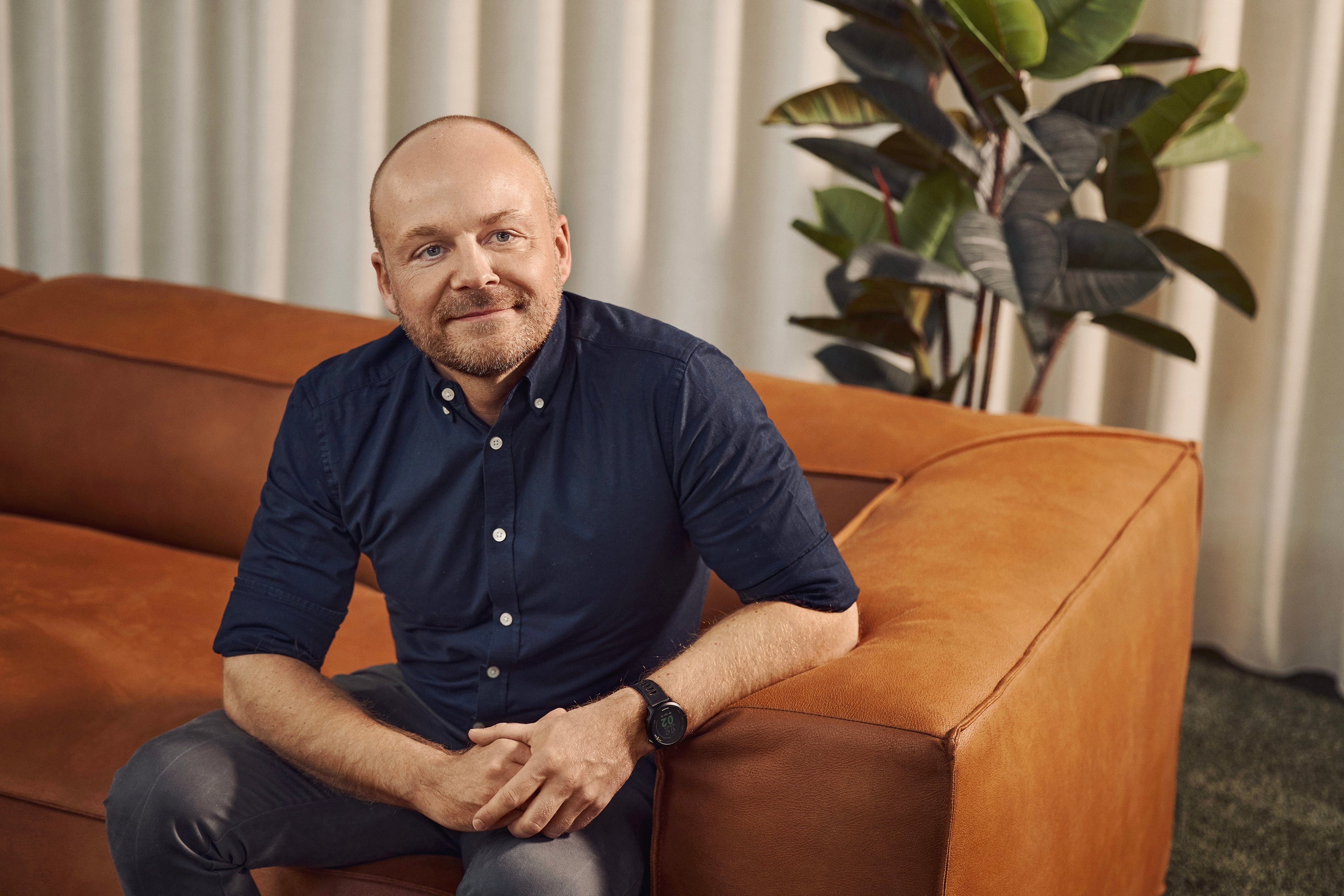 Man in navy shirt sitting on cognac-colored sofa in front of white curtains and green plant.